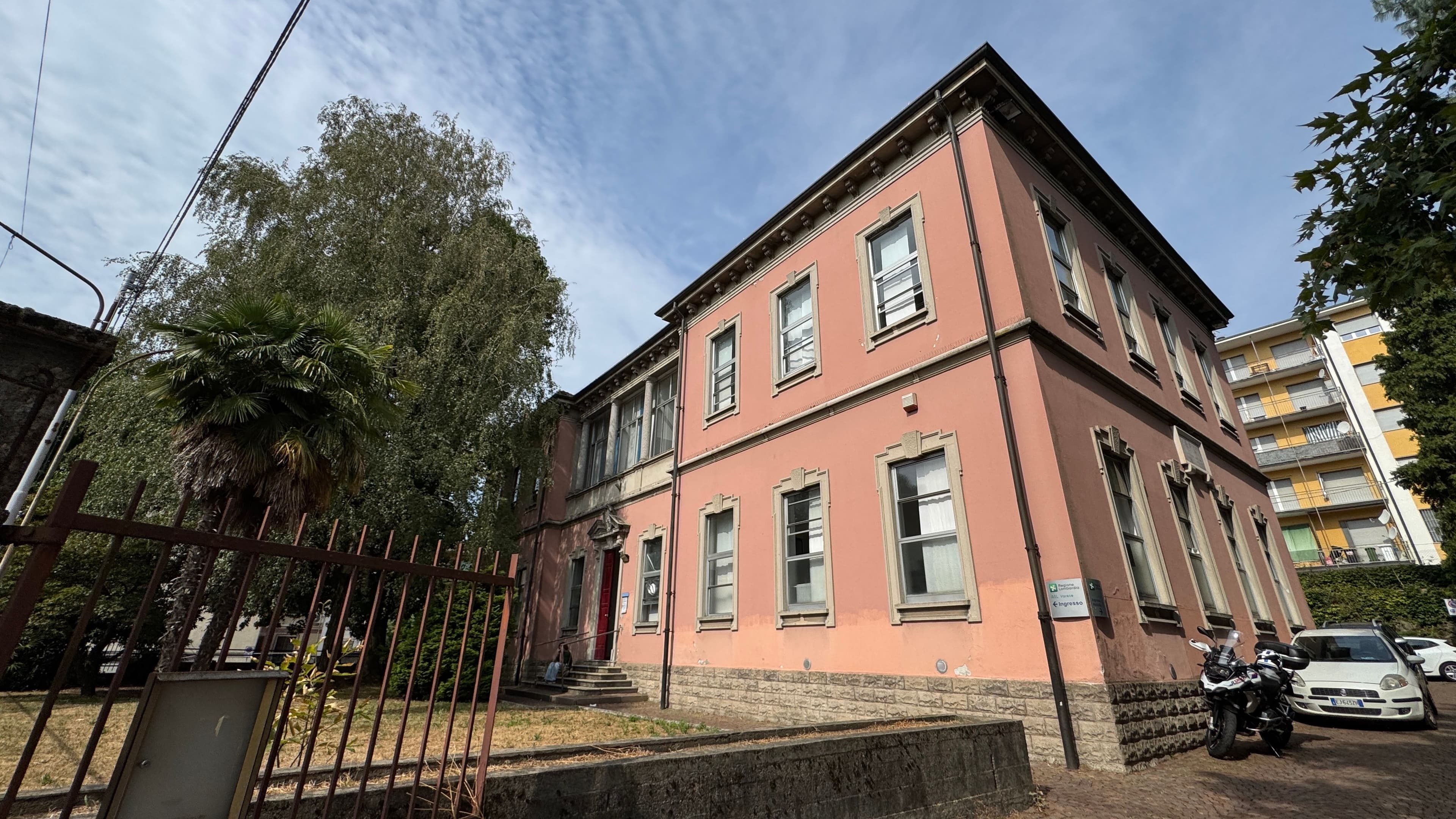 Panoramica dell’esterno della Casa di Comunità di Gazzada. L’immagine mostra un edificio storico con facciata rosa, ingresso principale, alberi nel giardino circostante e area di accesso alla struttura sanitaria
