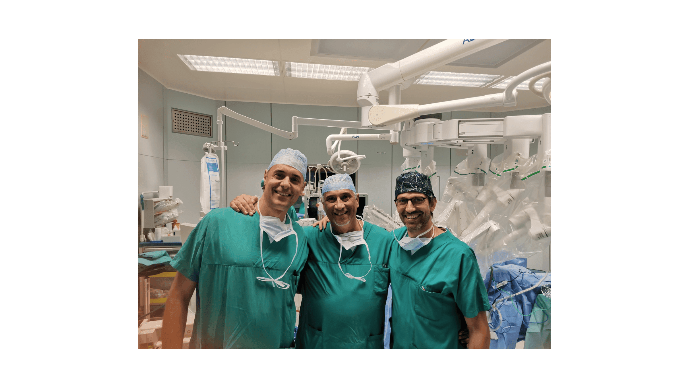Tre operatori sanitari in sala operatoria, sorridenti e in abbigliamento chirurgico verde, posano davanti a un sistema robotico utilizzato per gli interventi. Sullo sfondo sono visibili apparecchiature e strumentazione della sala operatoria.
