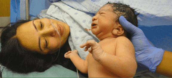 Madre sdraiata accanto al neonato subito dopo il parto, assistita da personale sanitario con guanti, in ambiente ospedaliero.
