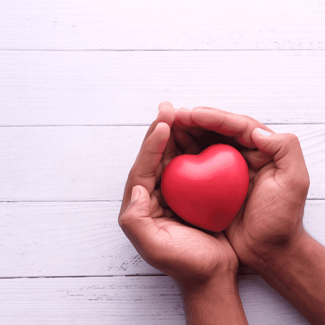 Mani aperte che proteggono un cuore rosso, simbolo di cura, solidarietà e donazione.
